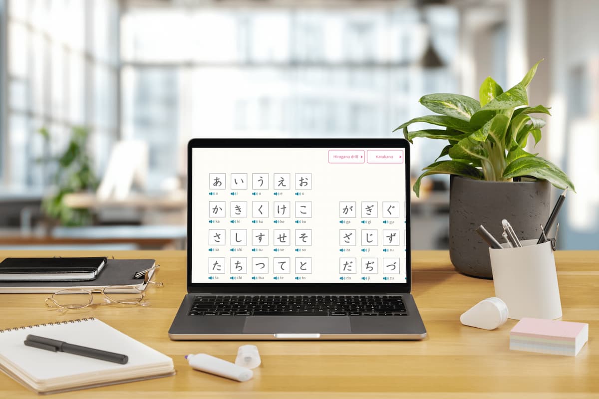 明亮現代的辦公空間內，木質桌上放置一台筆記型電腦，螢幕顯示日語平假名與濁音的學習介面，周圍擺有筆記本、眼鏡、文具與盆栽，呈現線上學習日語的情境。