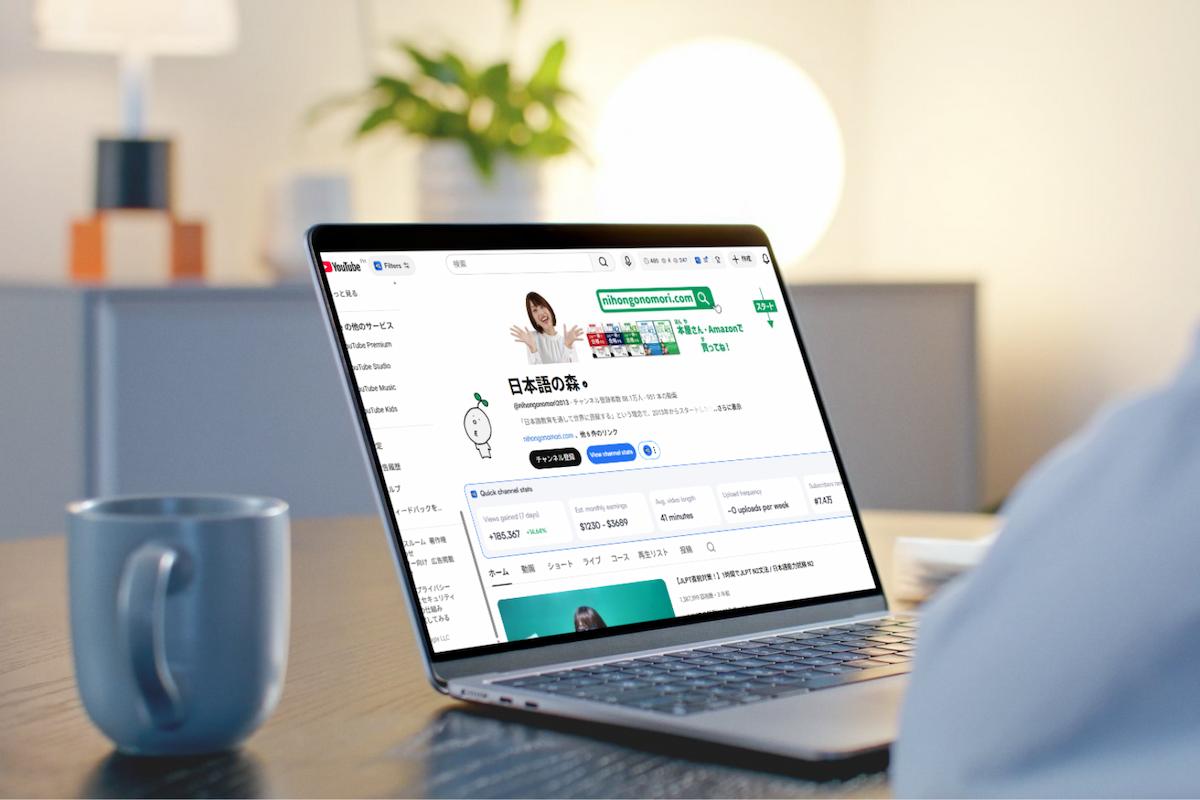 桌上放置一台筆記型電腦，螢幕顯示日語學習 YouTube 頻道「日本語の森」的頁面，旁邊有一個藍色馬克杯與檯燈，背景為溫馨明亮的室內空間，呈現居家線上學習日語的情境。