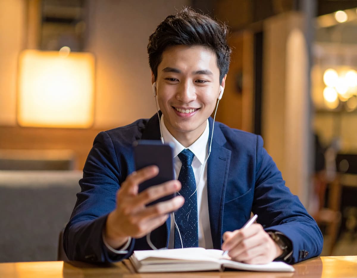 一名穿著西裝的年輕男子正在咖啡廳裡，佩戴耳機，微笑著看著手機。他的桌上有一本筆記本，他正準備寫筆記，顯示出他正在進行線上學習或通話。