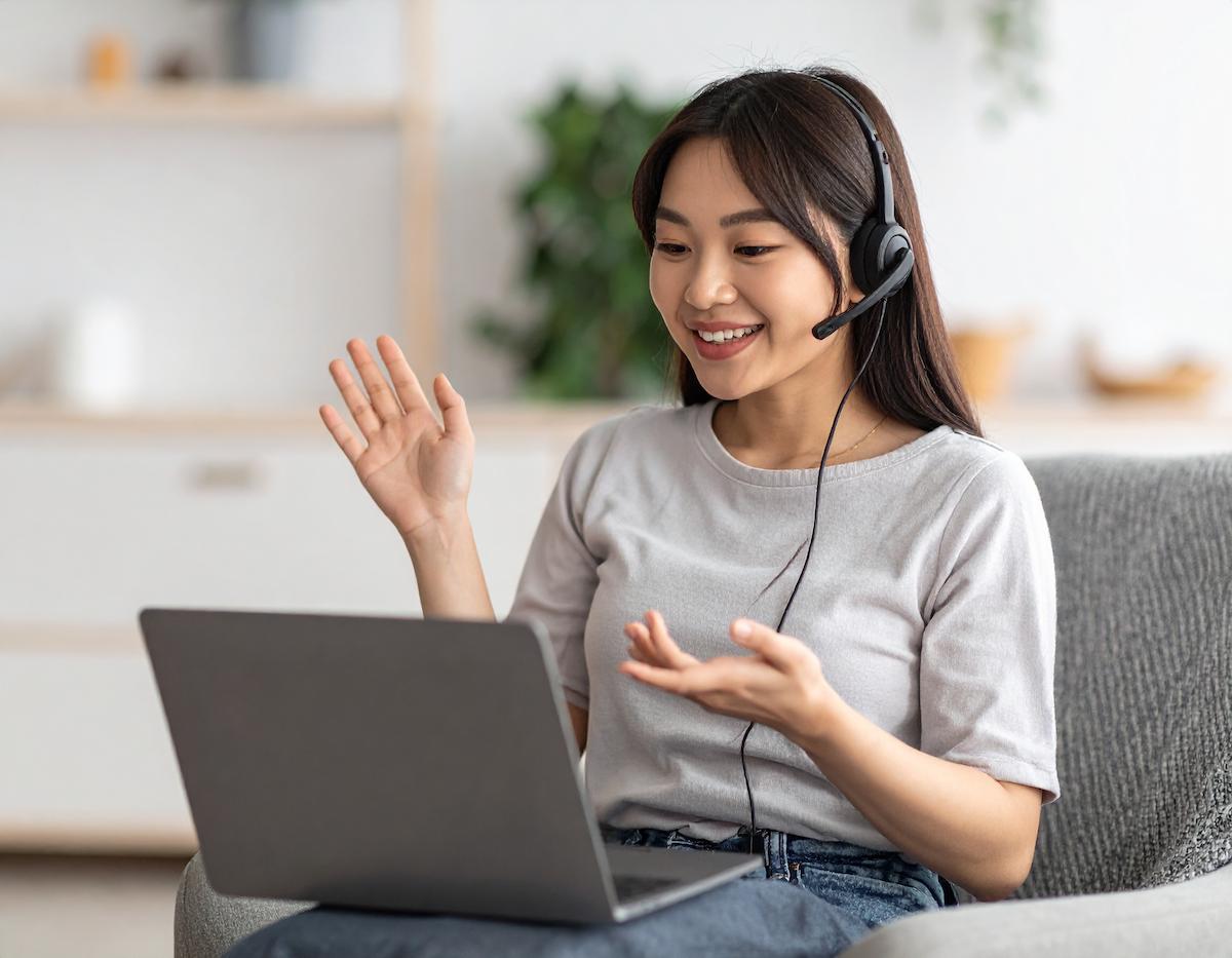 一位年輕女性戴著耳機麥克風，坐在沙發上對著筆電微笑交談。她一手揮手問候，一手做出說話的手勢，畫面呈現她正在進行線上課程或視訊通話，氛圍輕鬆愉快。